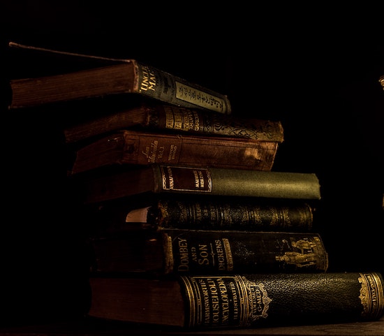 Aesthetic image for the post Delectable Dystopians to Read in 2021. There are 7 antique books stacked haphazardly on top of each other skewing to the left of the frame. They are all dark in nature along with the image as it is quite moody with a black background. Only the seven novels are in focus with lighting predominately on the right side.