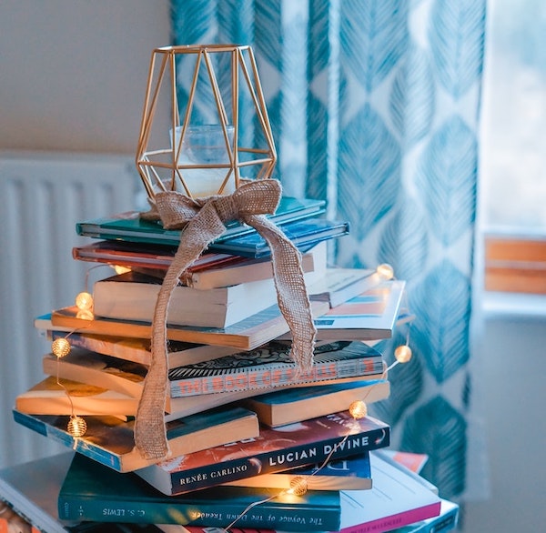 Aesthetic image for New 2021 Novels You Must Read | January - June Edition. This is a warm toned image with a haphazard stack of books in the forefront and a small lantern on top with a bow trailing onto the books. There are also string lights wrapped around the books. In the background, it is a white wall and a blue and white curtain.