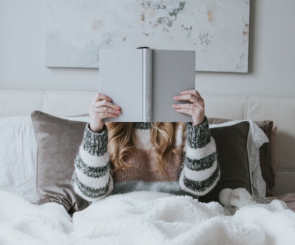 Aesthetic image for 8 fantastic novels from 2020 to read in 2021. It is a bright image with a white overtone. There is a woman sitting up in her bed holding up a book. The book is grey and covers her face. A fluffy white blanket is draped over her and covers the lower half of the image, her sweater is white and grey striped, and the pillow she is resting on is brown.