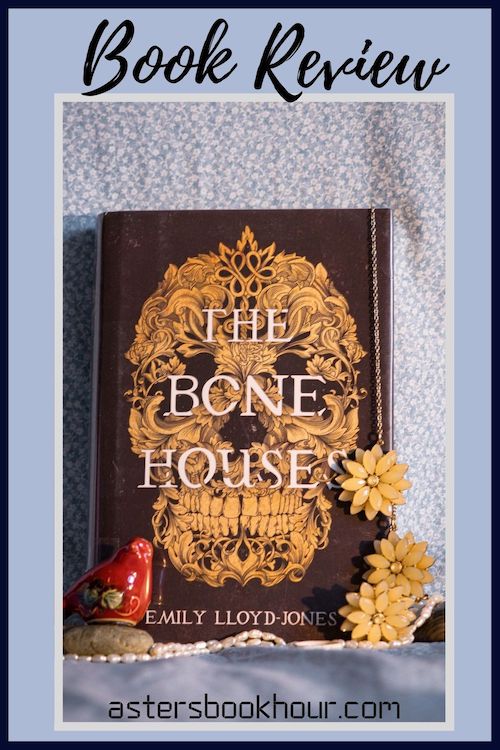 The pinterest image for The Bone Houses by Emily Lloyd-Jones book review. There is a blue floral print background with the novel centered in the middle and the cover facing the front.
